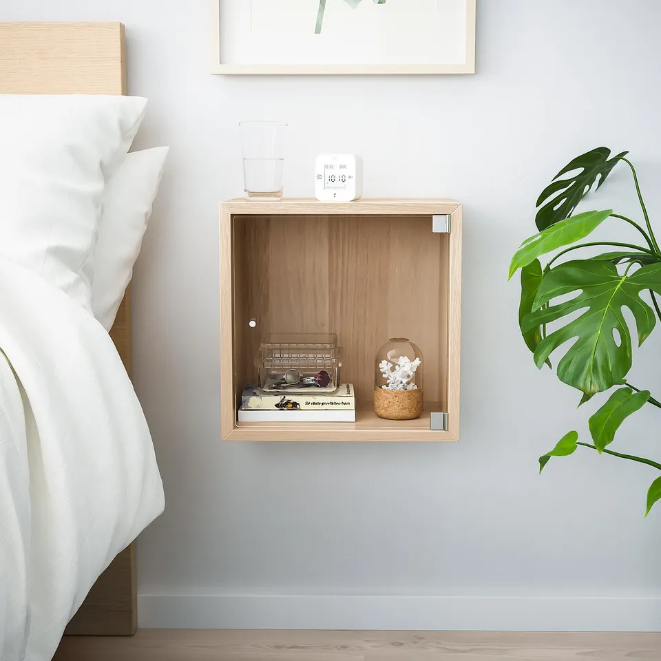 Wooden EKET shelf with decor and plant beside a bed.