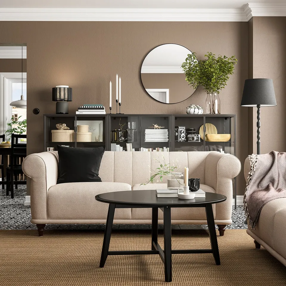 Modern living room with beige sofa, black coffee table, dark bookshelf, plants, candles, and round mirror.