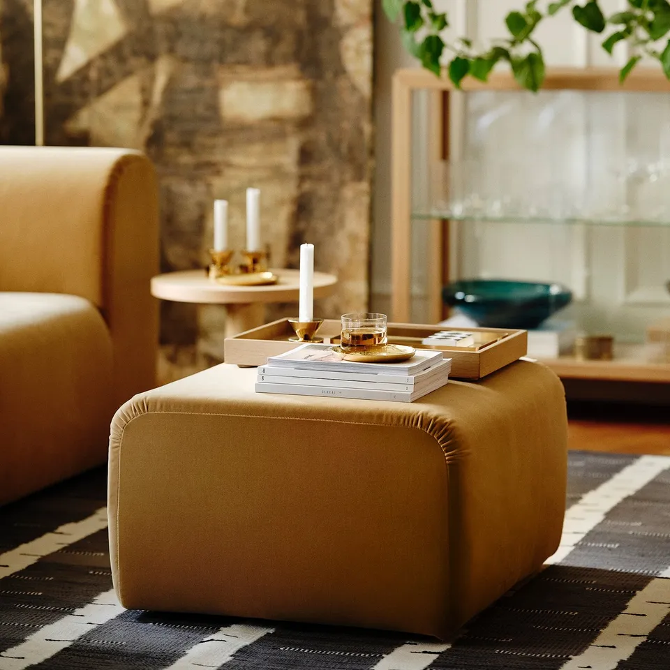Small, golden-brown, square ottoman on striped rug near large sofa, holding books and tray with candles, in cosy living room.