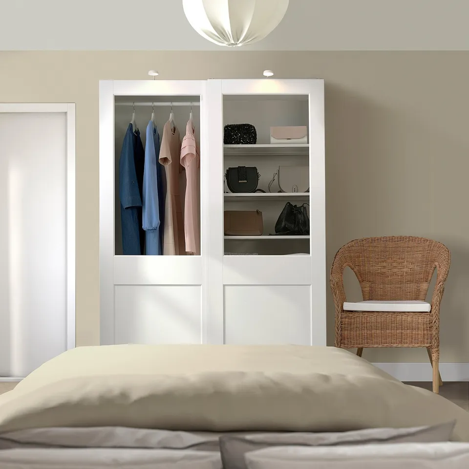 Minimalist bedroom with large beige bed, wicker chair, spacious white wardrobe displaying clothes and accessories.