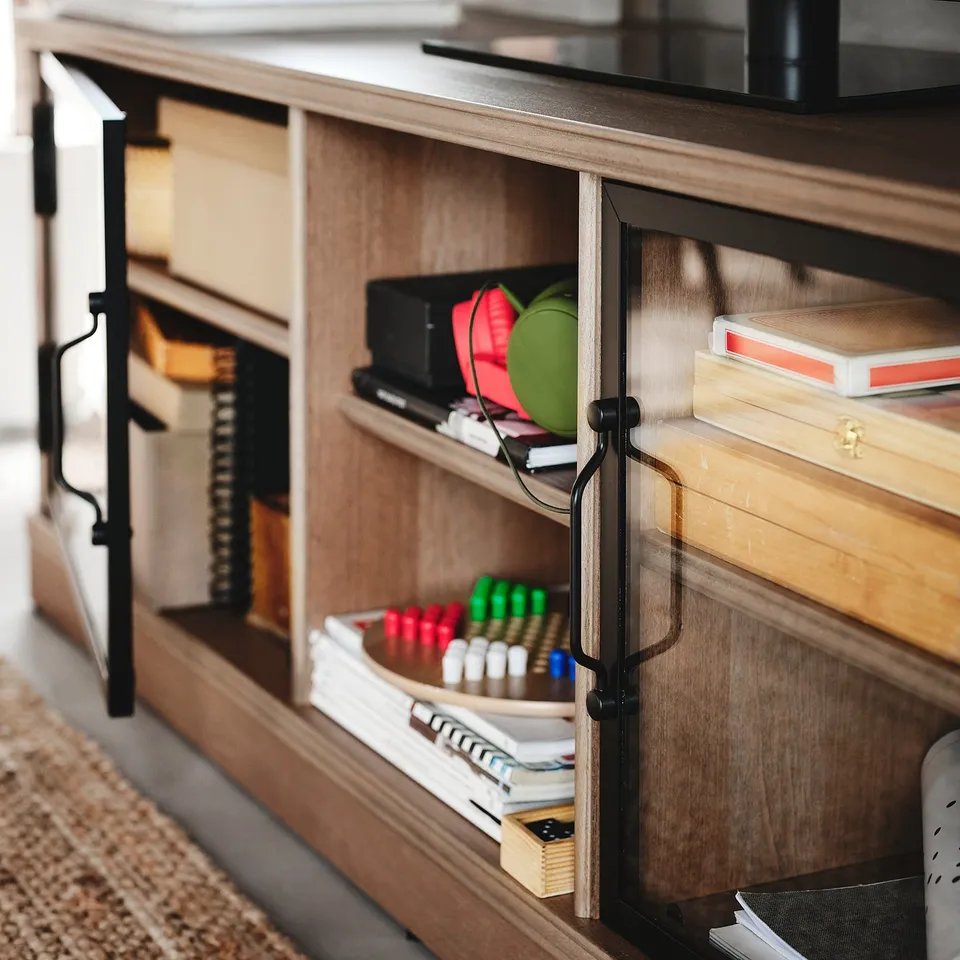 Wooden tv cabinet with glass doors, storing various items. Dark handles, natural wood finish.