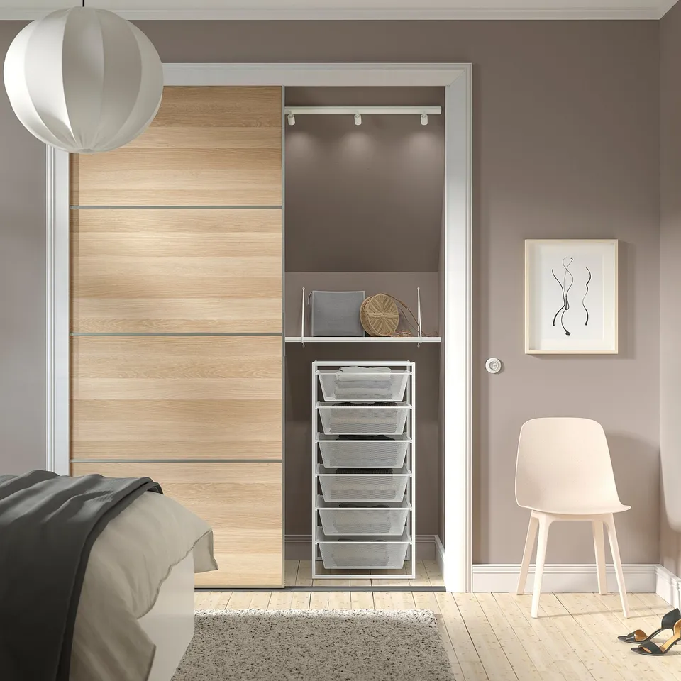Modern bedroom with large lightwood JONAXEL storage unit, including metal mesh baskets, near bed. White pendant light and chair, grey walls.
