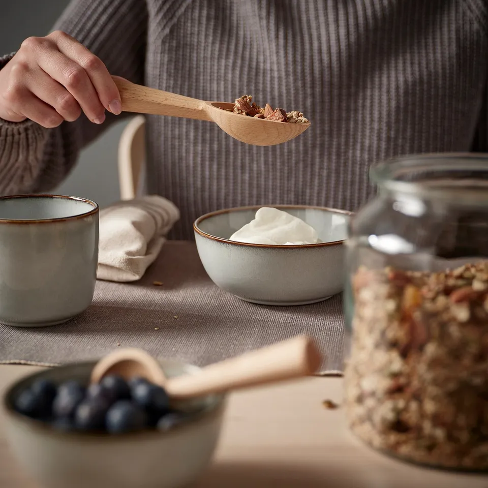 A person holding a wooden spoon filled with granola above a bowl of yoghurt. The table has berries and a jar of granola.