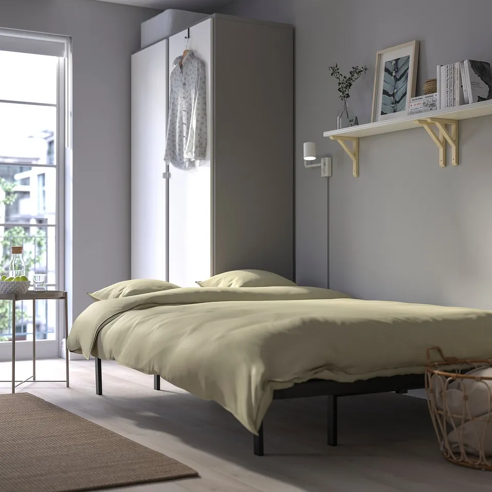 Modern bedroom with beige bed, hanging garment, shelf with books and decor, and large window.