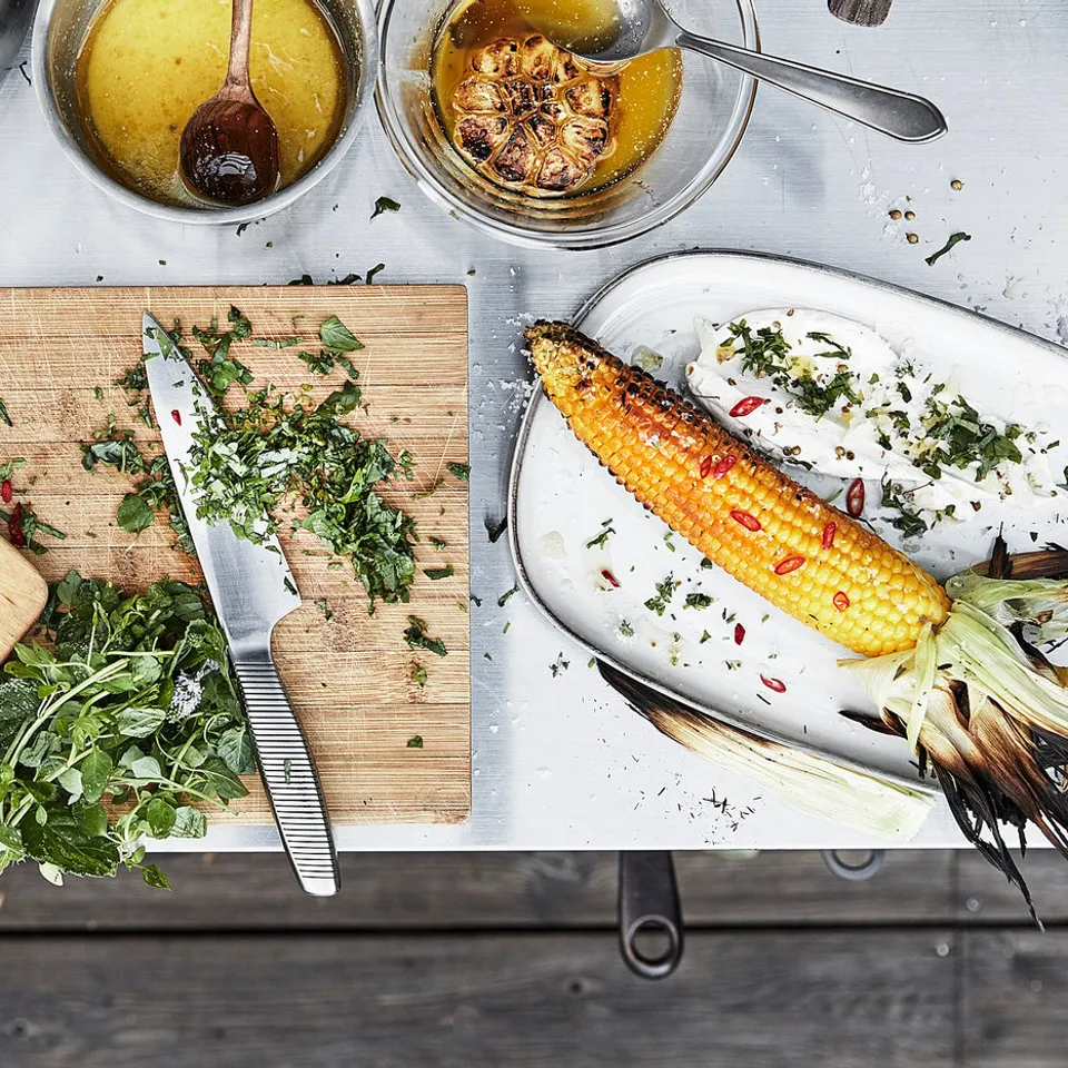Grillske̊r prep: cutting board, knives, herbs, chilli, corn on steel/wood island.