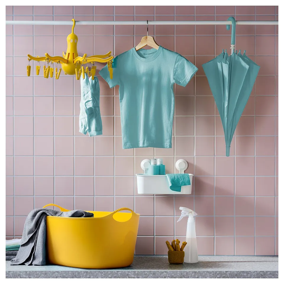 Bright yellow PRESSA dryer hangs on pink tiled wall, holding blue shirt & socks. Yellow basket & spray bottle below.
