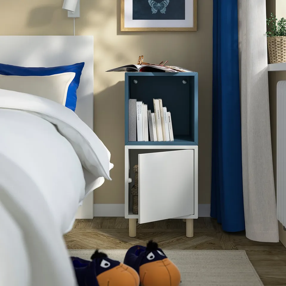Small white & blue EKET storage unit beside a bed. Holds books on top and has a cabinet below. Suits compact spaces.