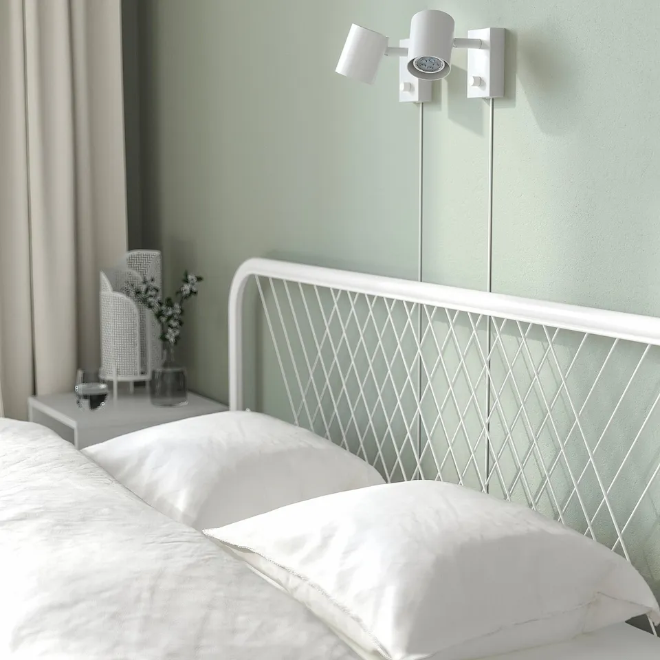 Modern white bed with net-patterned headboard, two adjustable wall lamps beside it, and a small bedside table with a vase and ceramic lamp.