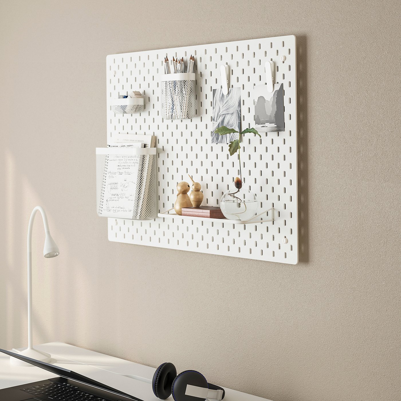 A white pegboard named SKÅDIS organises items like scissors, notes, and a vase on a wall above a desk with a lamp and laptop.