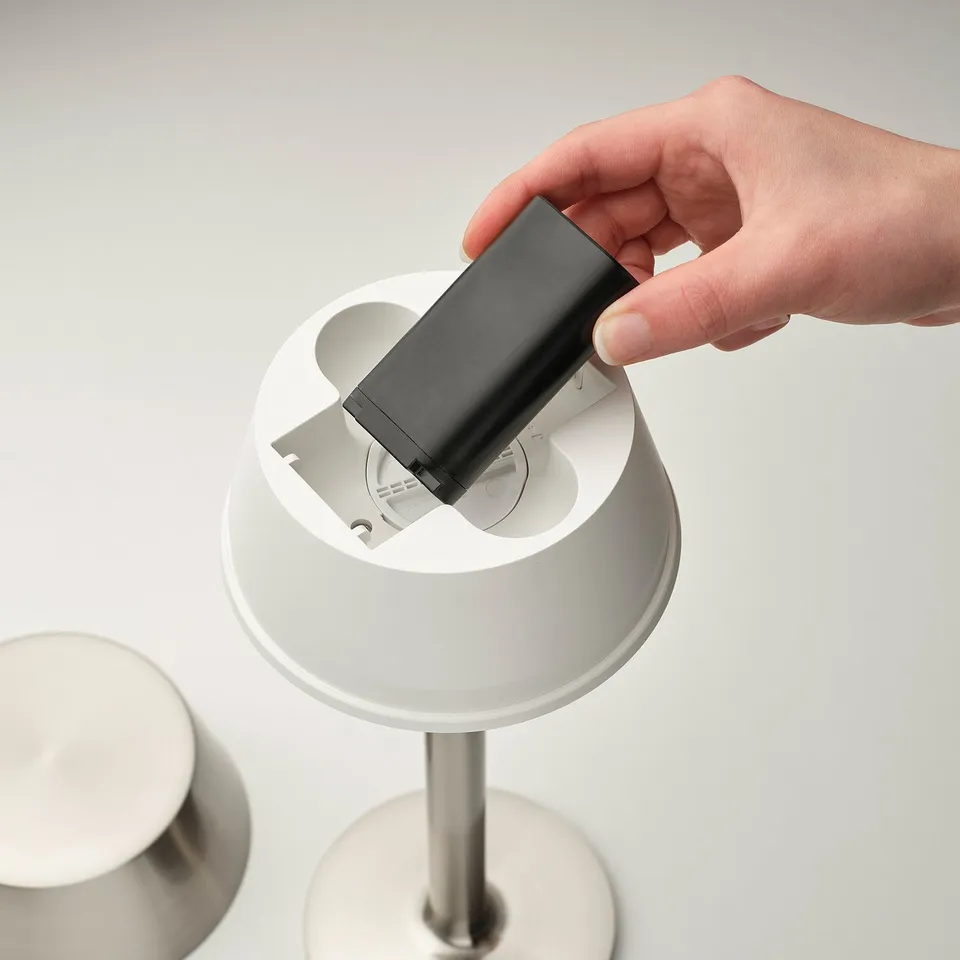 Hand placing battery into AVHÅLL portable lamp, showcasing its easy, wireless setup.