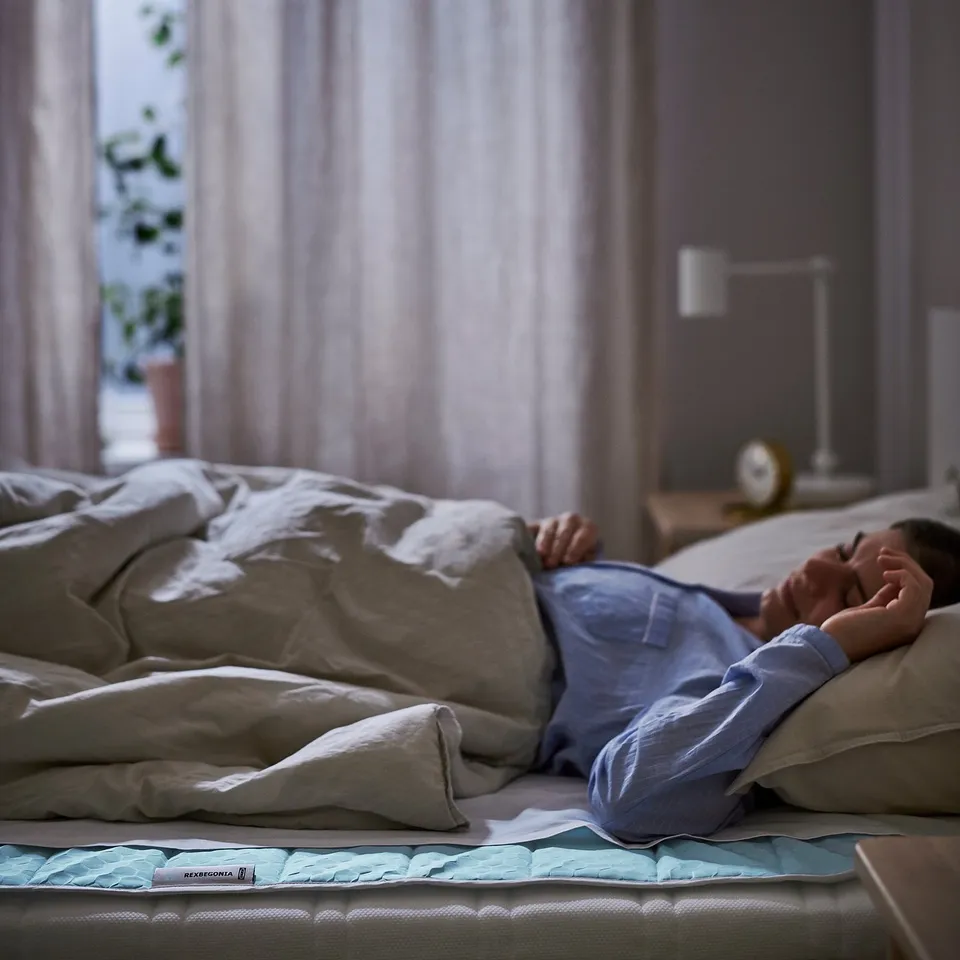 A person resting on a bed with a beige REXBEGONIA cooling mattress protector, which helps regulate temperature and protect the mattress.