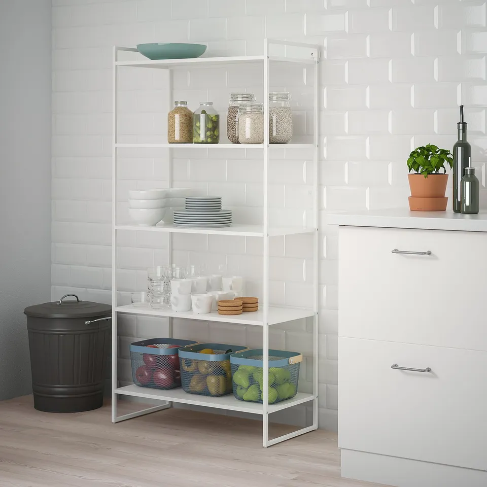 JONAXEL white metal shelf unit holds kitchen items, including jars, bowls, plates, and fruits in baskets, fitting neatly beside counter and cabinet.