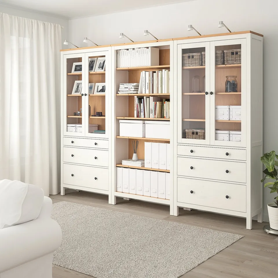White HEMNES unit with glass doors & drawers, holding books & decor, minimalist & room-dominating.