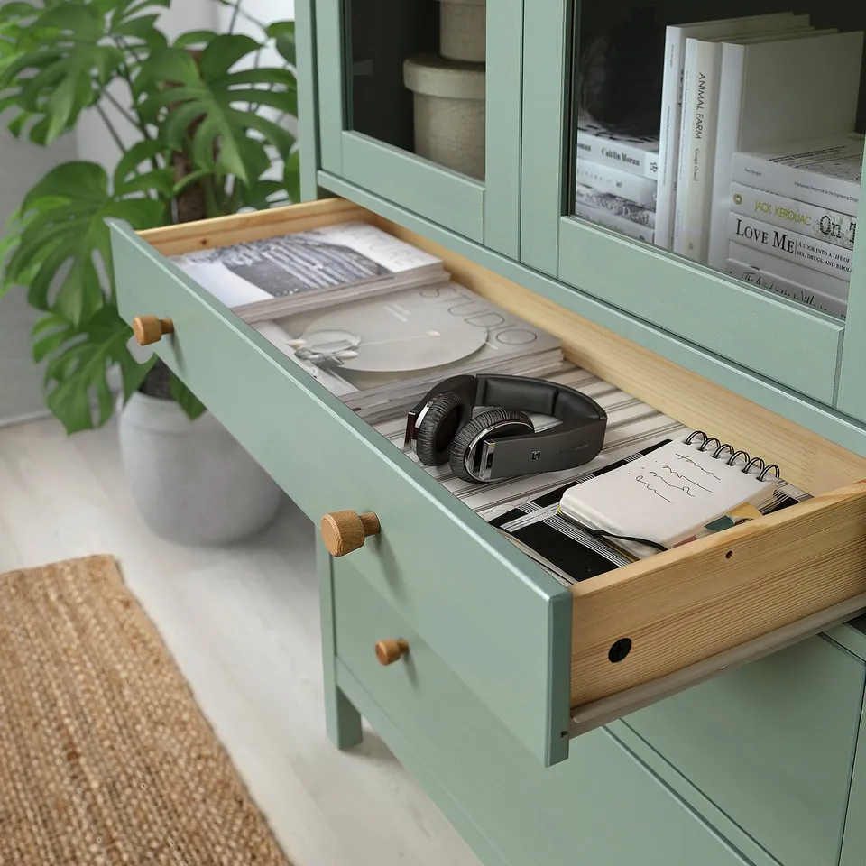 Pale green cabinet with open drawer showing headphones, notebooks, and records. Plant and books on shelves.