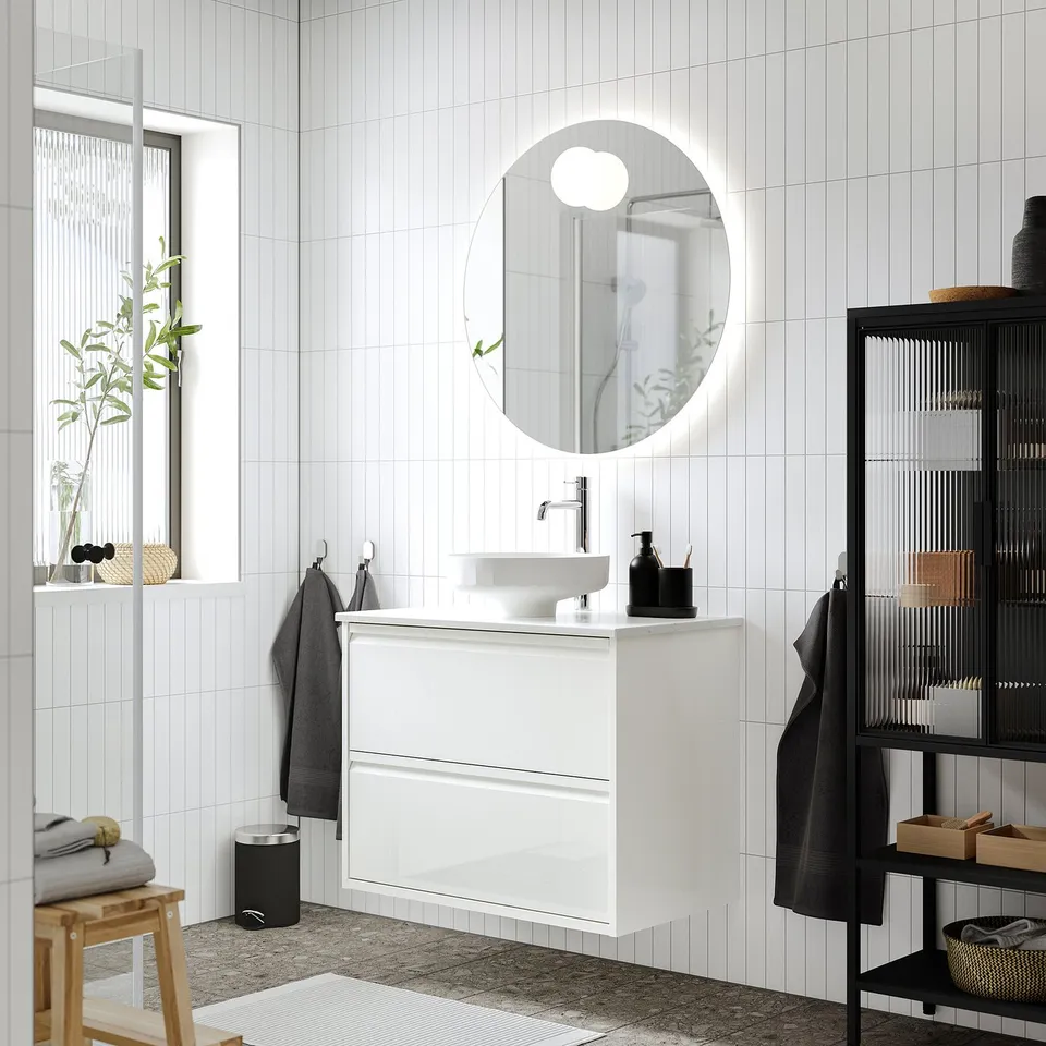 Modern white bathroom with sleek cabinet, round sink, large mirror, dark storage, and wooden stool with plant.