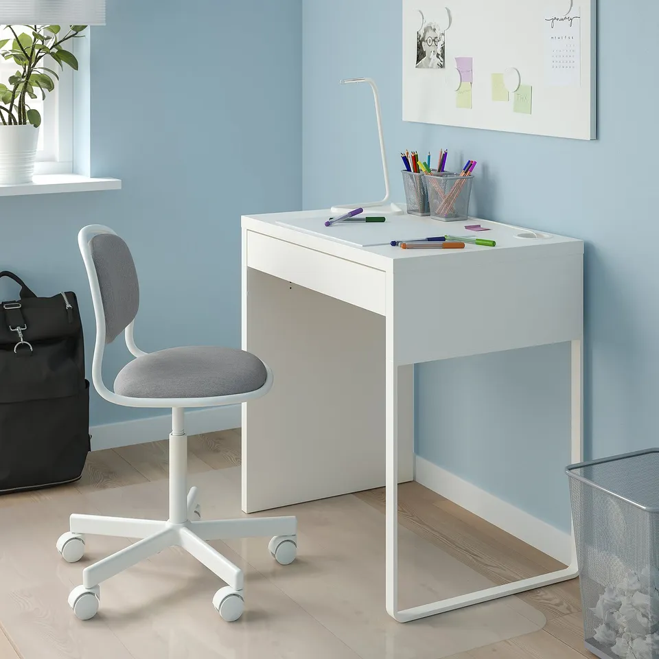Small white kids study desk and chair. Bright blue wall, pens, lamp, plant, and bag beside.