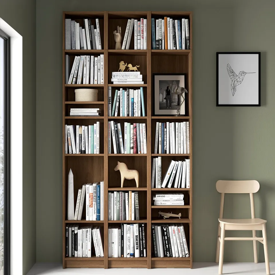 Large wooden BILLY bookcase against green wall, packed with books and decorated with small items. Accompanied by light wooden chair.