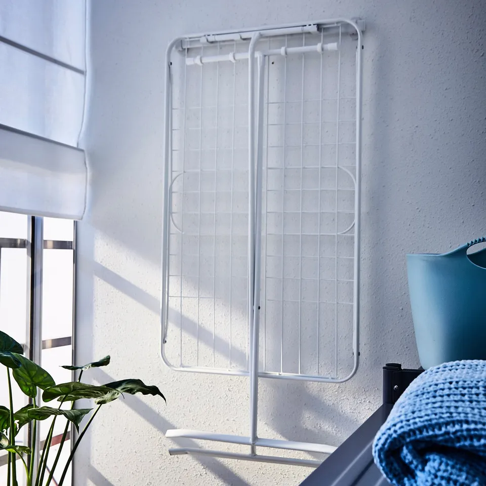 White MULIG drying rack with foldable wings against wall, taller than window, flanked by green plant and blue vase with blanket.