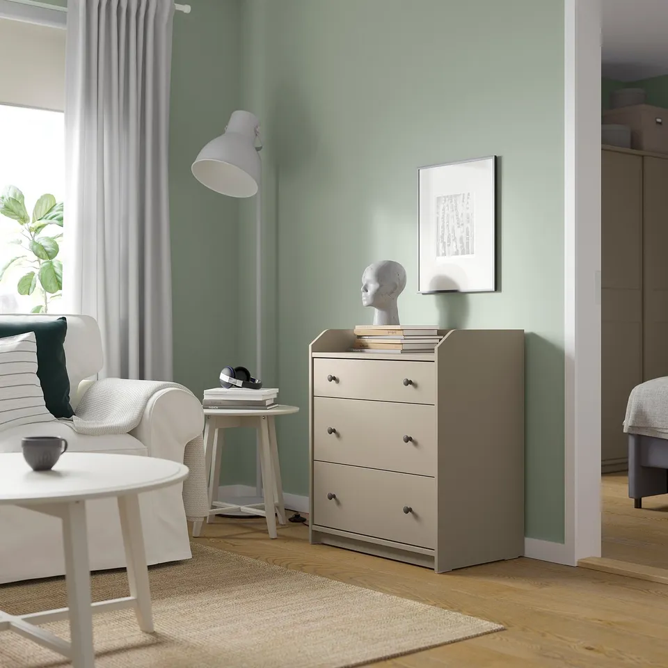 Modern living room with light green walls, a white chair, wooden floor, and a beige HAUGA chest of drawers with three drawers and a white lamp on top.