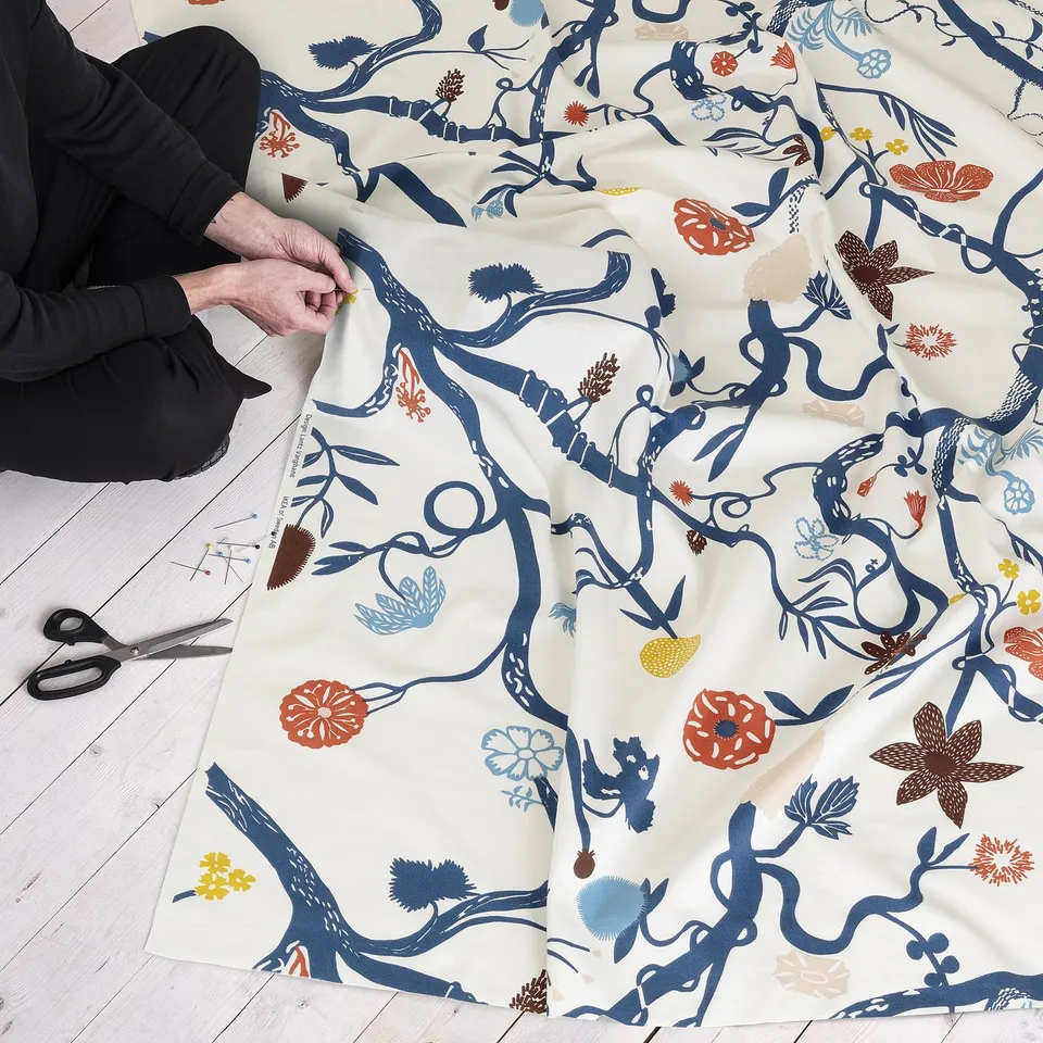 Person holding patterned fabric, scissors nearby. The fabric has blue branches, colourful flowers, and is laid out on the floor.