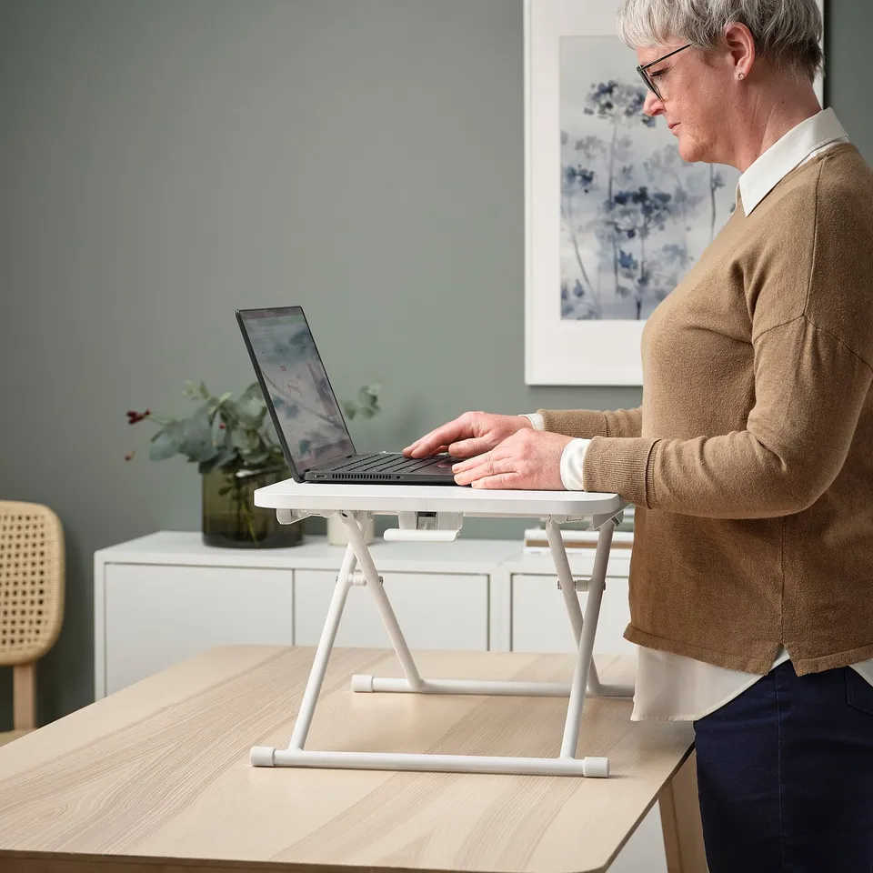 Person uses DUBBLA laptop stand, switching between sitting and standing. The stand elevates the laptop to eye level, promoting ergonomic posture.