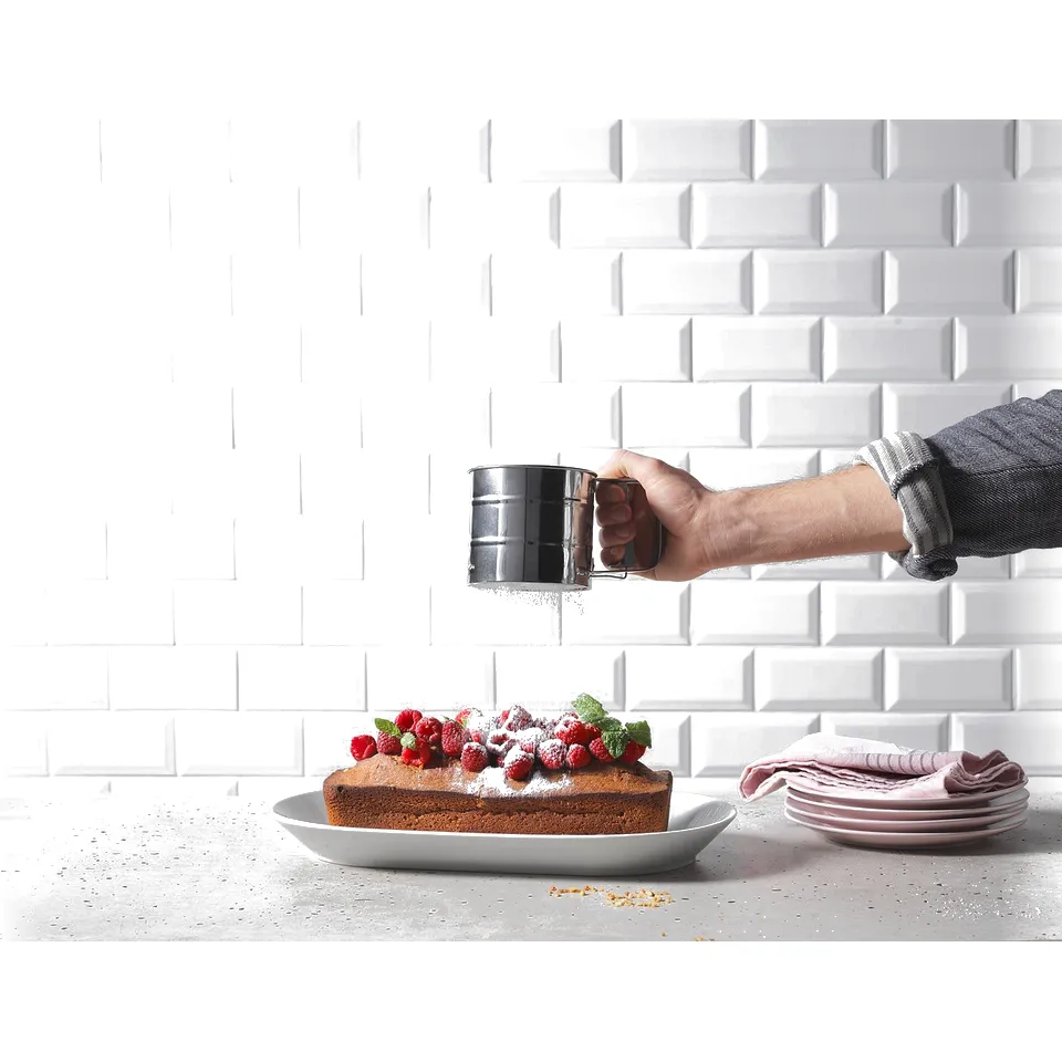 A person sprinkles icing sugar on a cake topped with raspberries and mint, placed on a white counter against a white tiled wall.
