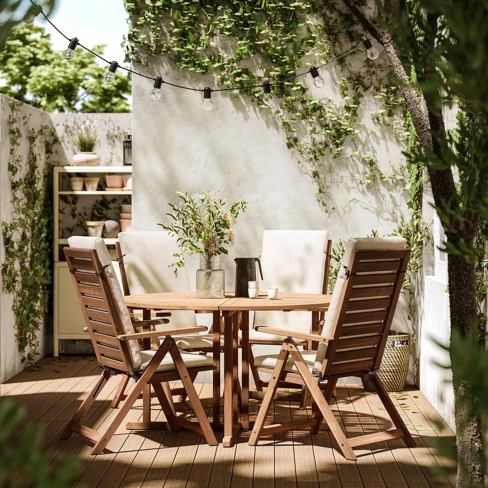 Outdoor patio set with wooden table, four matching chairs, and string lights overhead. Chairs have beige cushions.