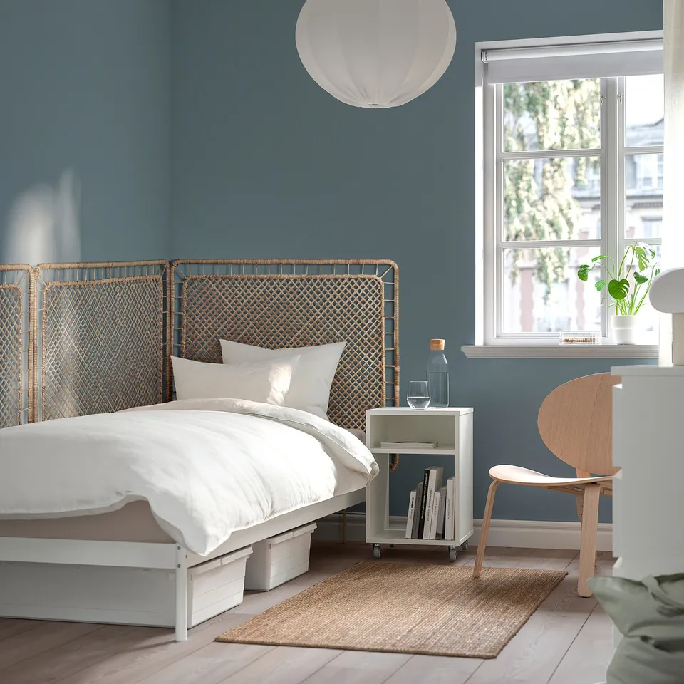 Blue bedroom with wicker headboard, white bed, side table with books and lamp, chair, jute rug.
