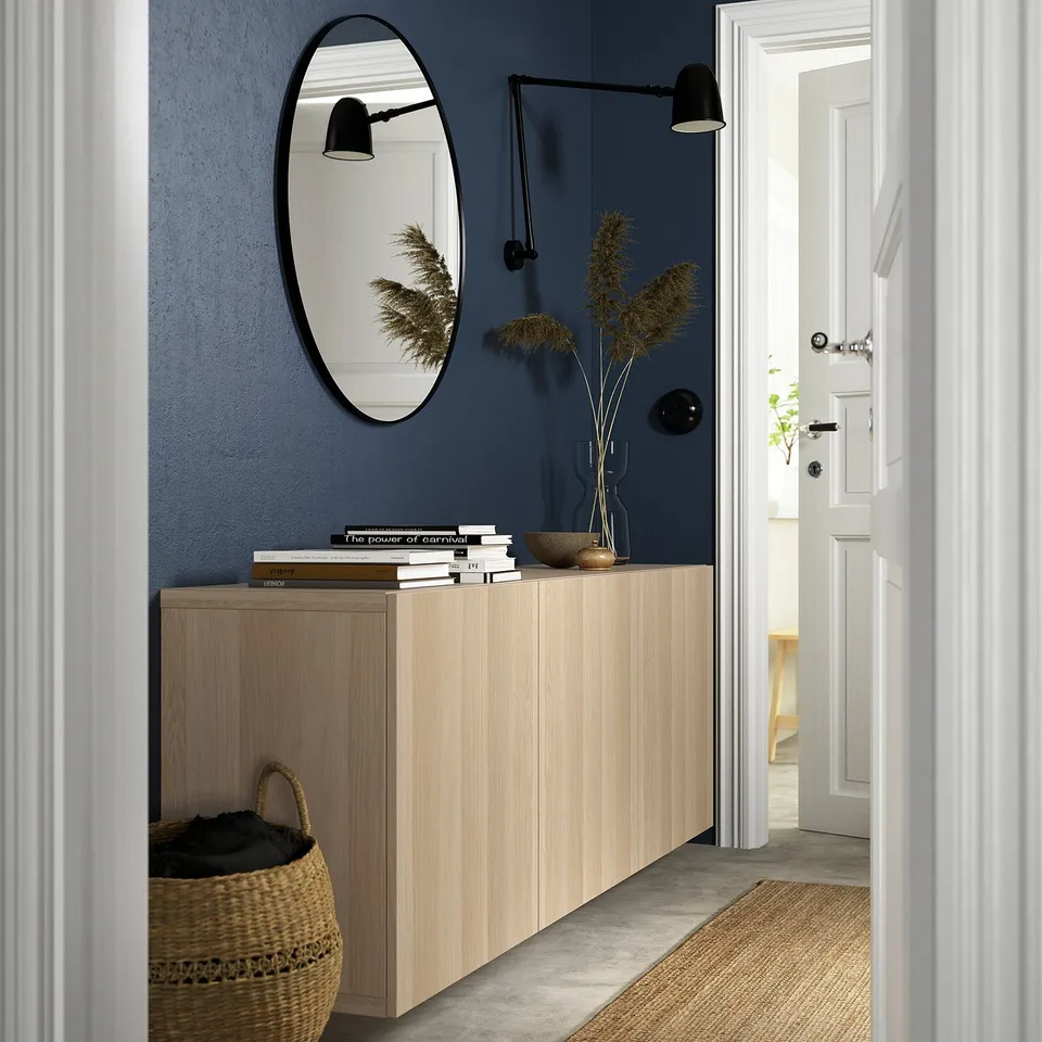 Modern entrance with LAPPVIKEN light oak cabinet, large mirror, wall lamps, dried plants, books, and basket. Blue wall contrast, white door.