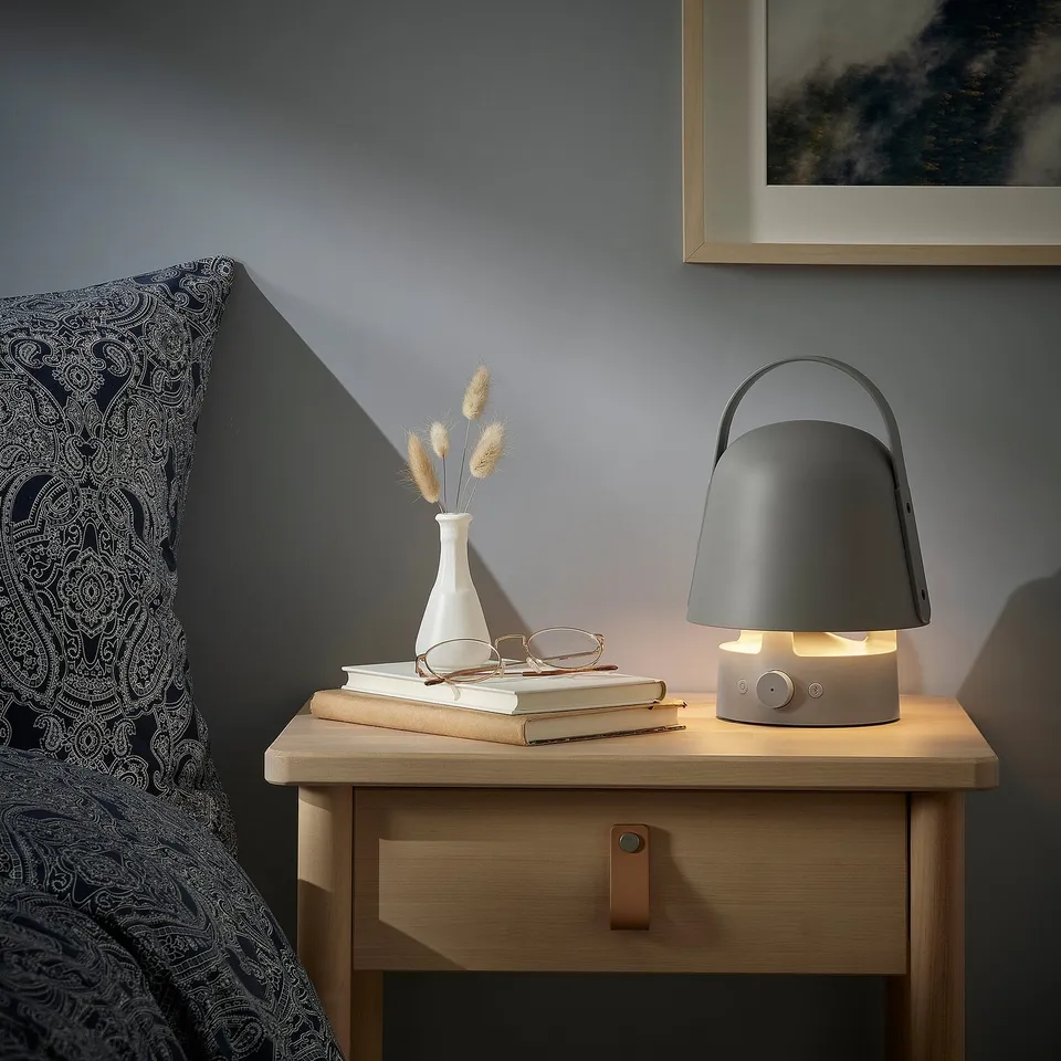 Grey VAPPEBY speaker lamp on wooden bedside table. Warm glow, books, glasses, dried plants. Cosy, organised.