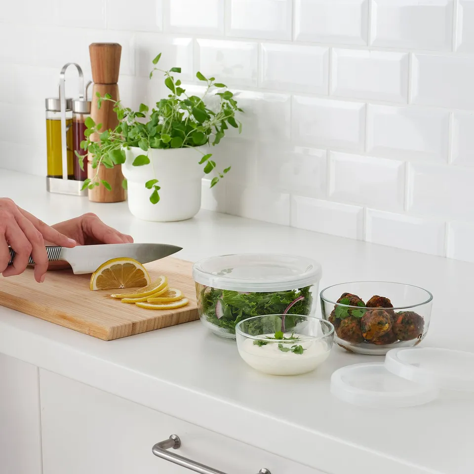 Hand slicing lemon on cutting board near glass containers with herbs and food on white kitchen counter.