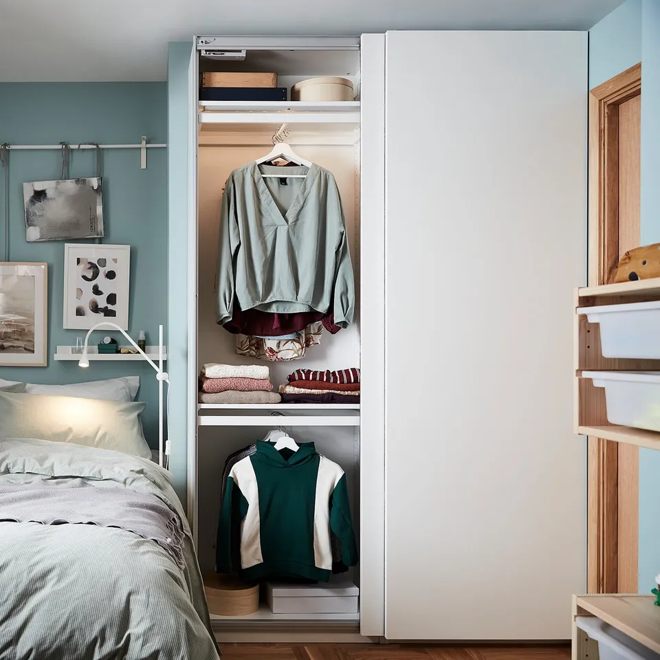 A modern bedroom a PAX wardrobe in white, holding blue and green clothes, with a bed and artwork.
