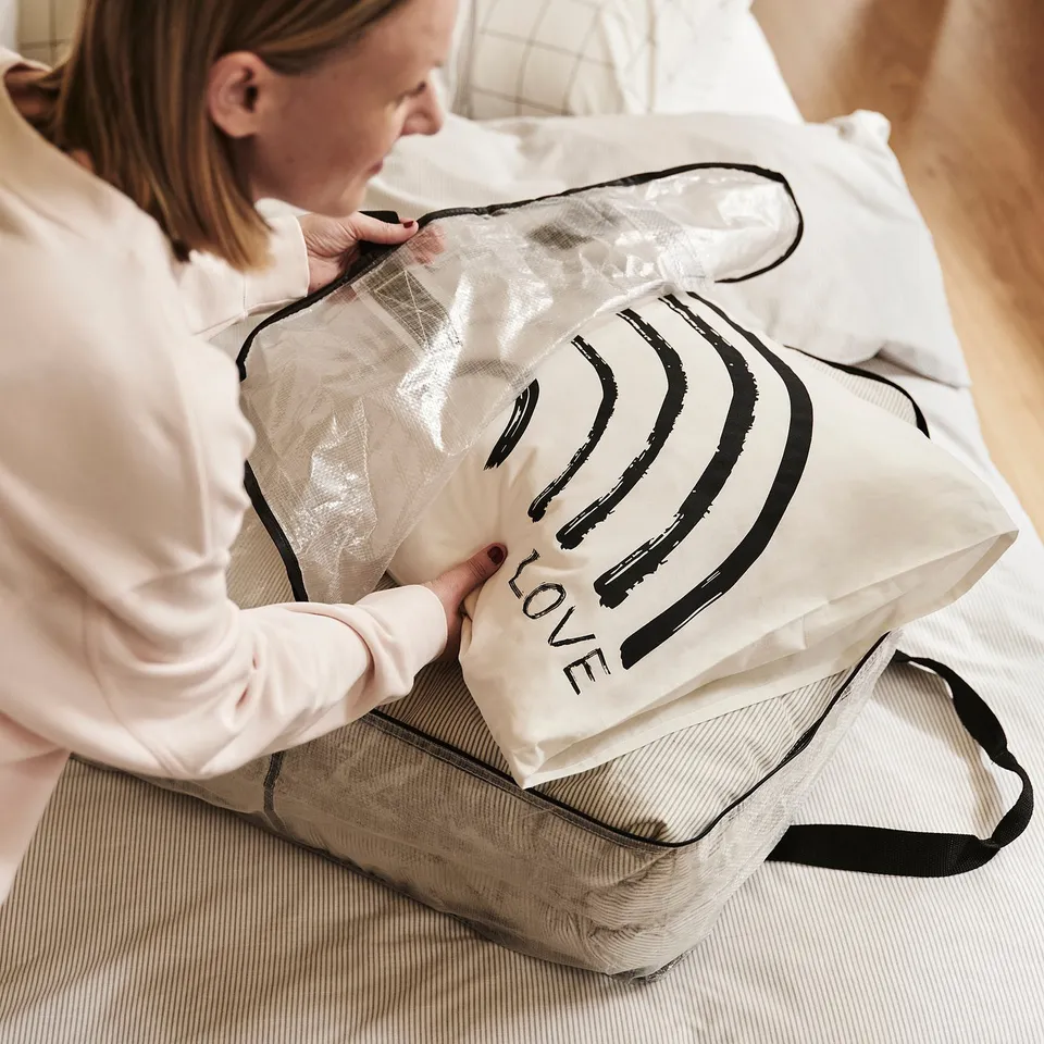 A person unfolds a large, durable DIMPA storage bag on a bed. The bag, labelled love, is transparent and designed for storing various items.
