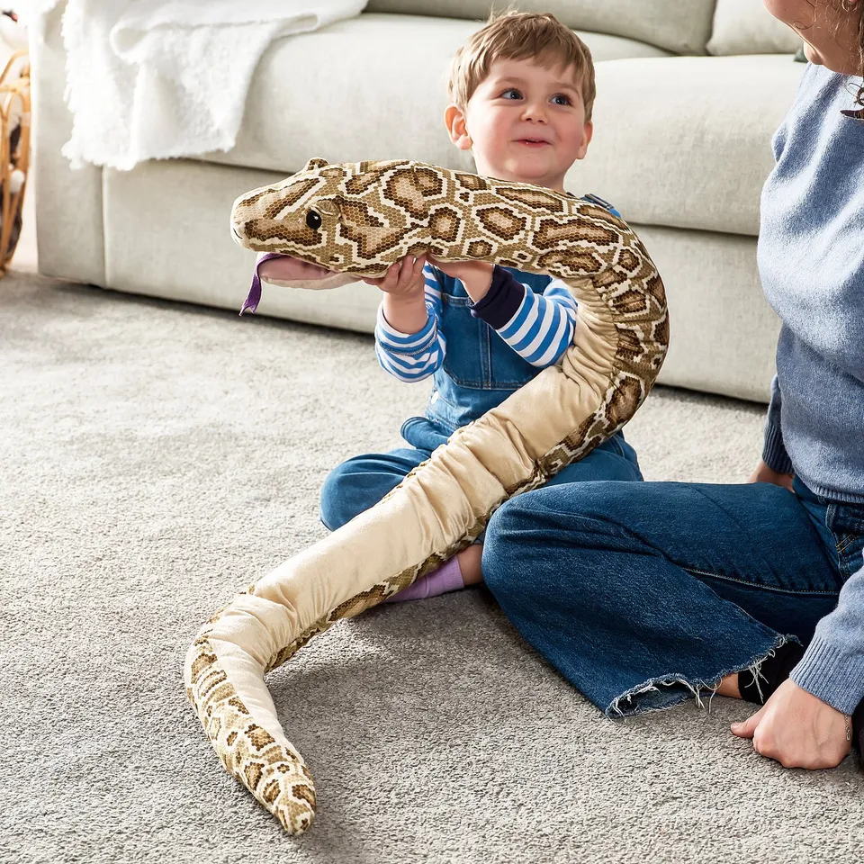 A child sits on carpet, holding a plush python toy. The toy speaks when a hand is placed in its head pocket.
