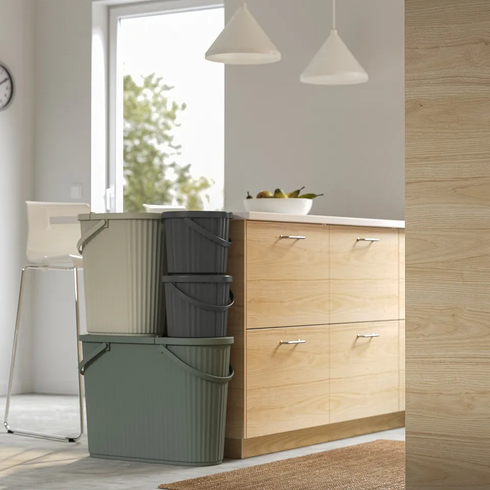 Stacked DAMMÄNG bins near a wooden cabinet, holding small waste items. Bins are designed with foldable handles for easy carrying.