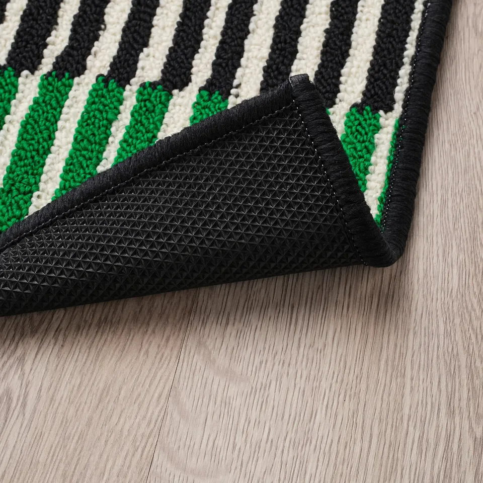 Striped door mat with black, green, and white patterns. Rubber backing for grip, placed on wooden floor.