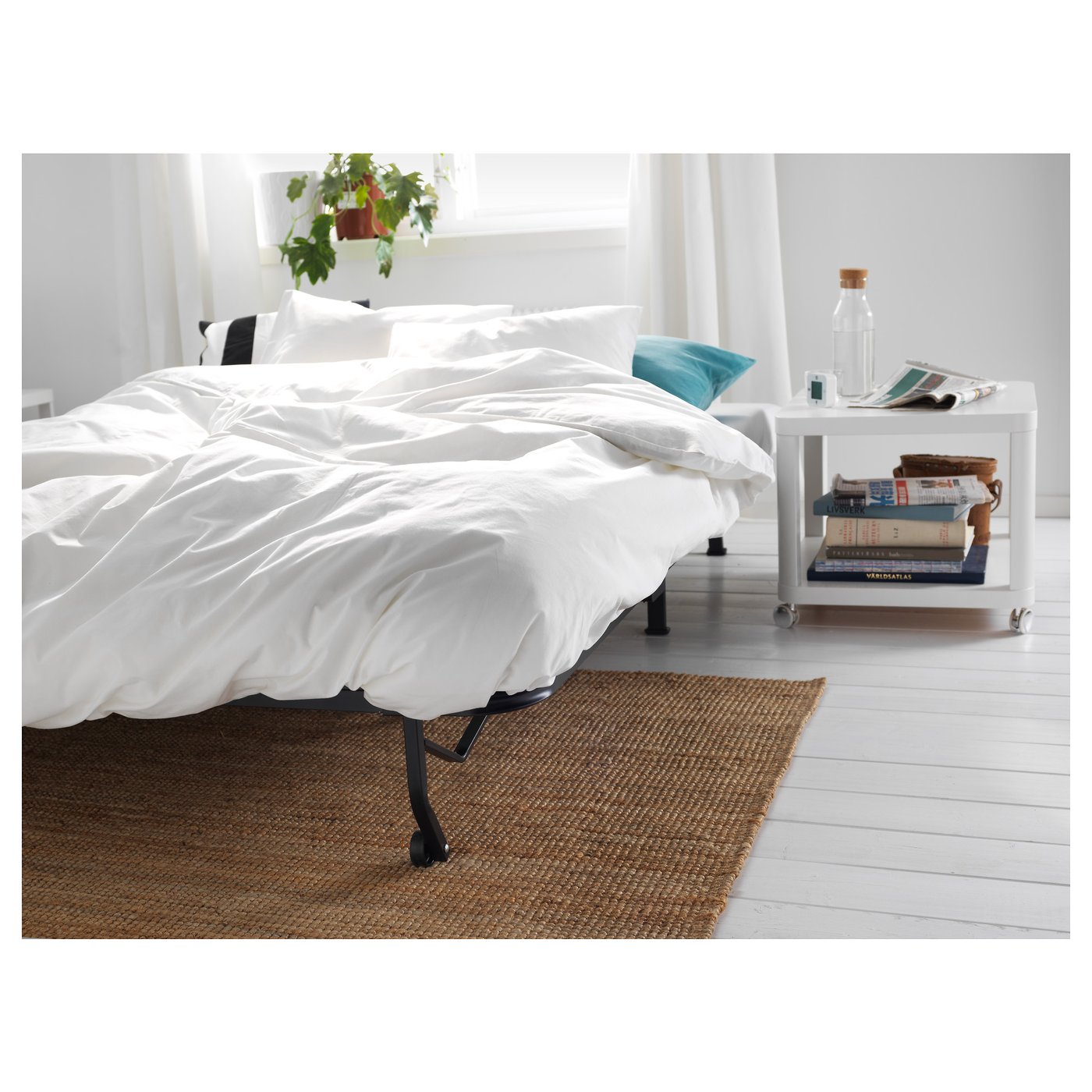 White sofa-bed, window, books, water bottle, blue pillow, rug.