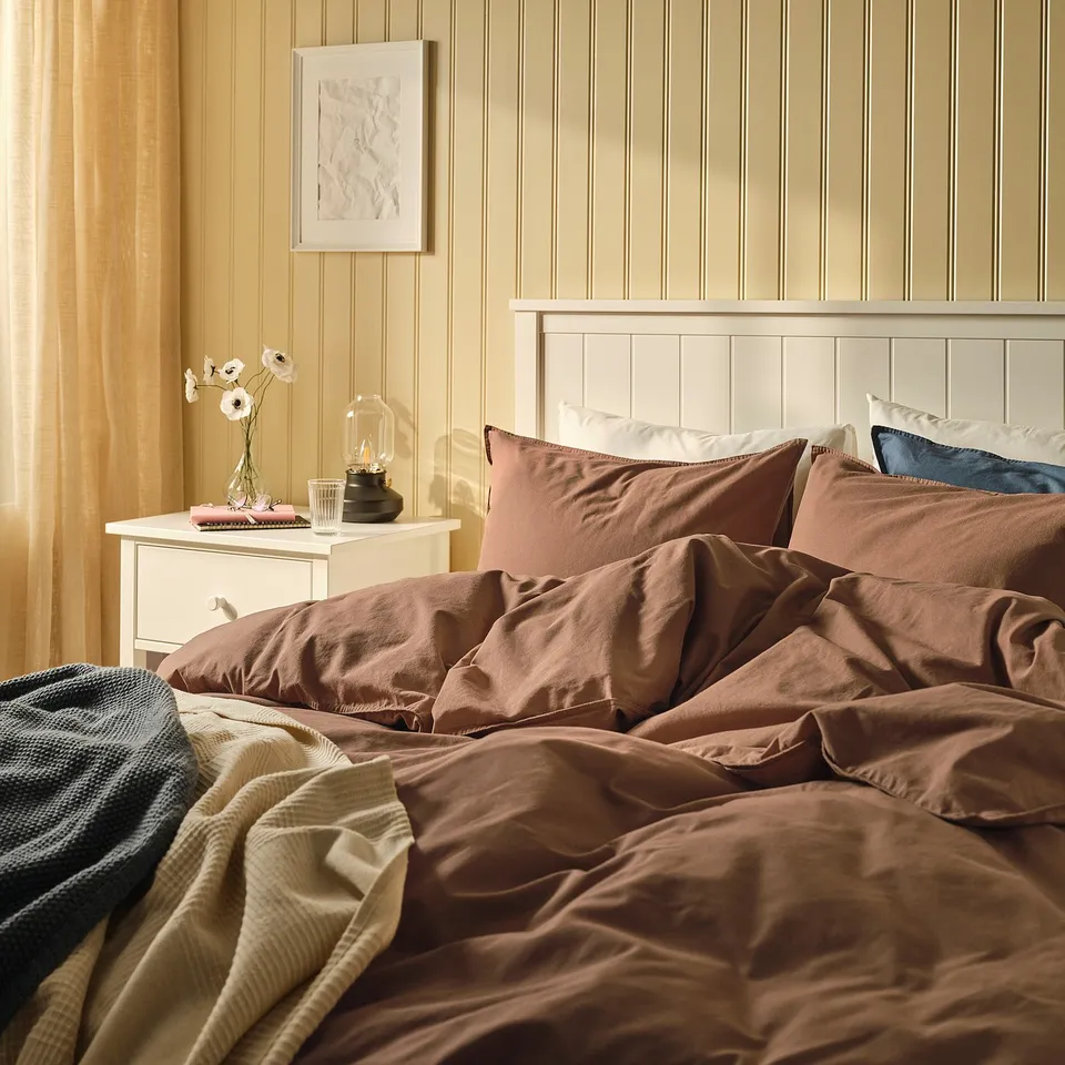Warm, cosy bedroom with ÄNGSLILJA duvet, wooden headboard, pillows, and bedside flowers.