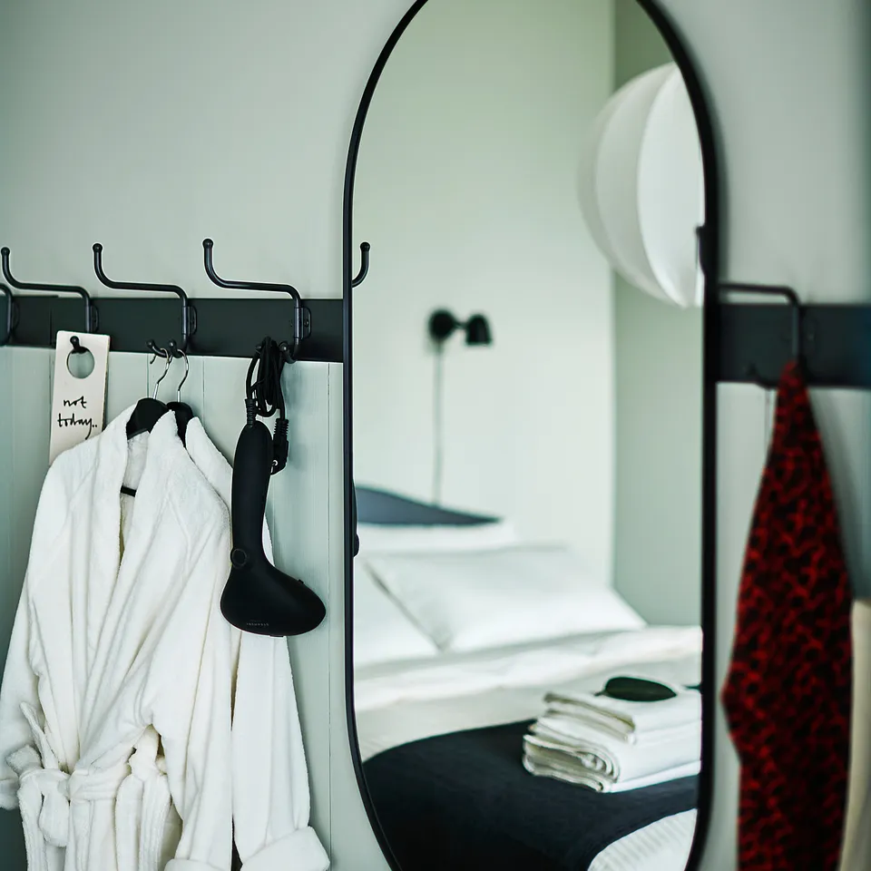 Large black-framed mirror hangs on a green wall, reflecting a white bed and stacks of towels. White robe and black shoe hangers flank it.