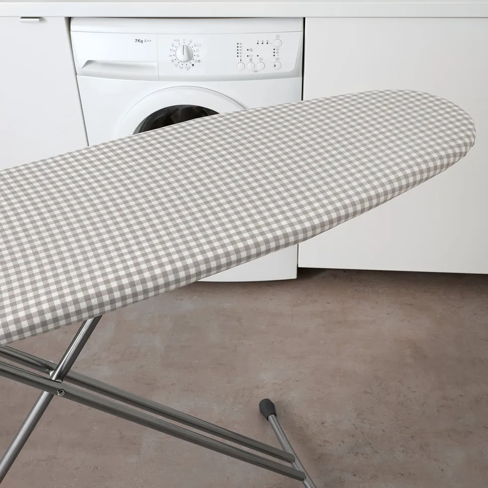 An ironing board with a chequered cover, padded for smooth ironing, stands in front of a washing machine.