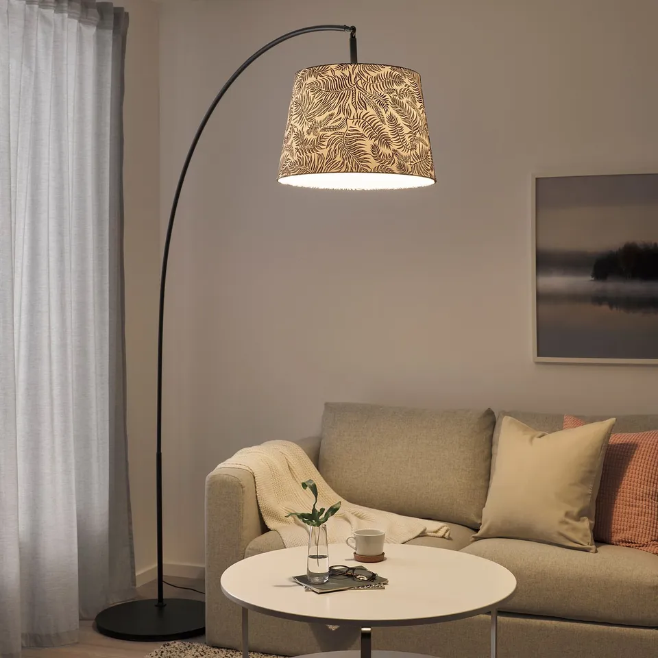Modern living room with beige couch, coffee table, and SKAFTET floor lamp arched overhead, featuring a patterned lampshade.