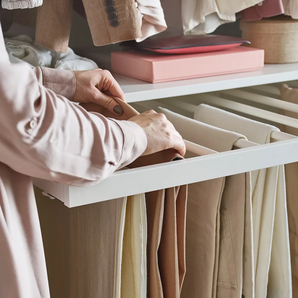 A person organises beige pants on a white KOMPLEMENT trouser hanger in a closet.