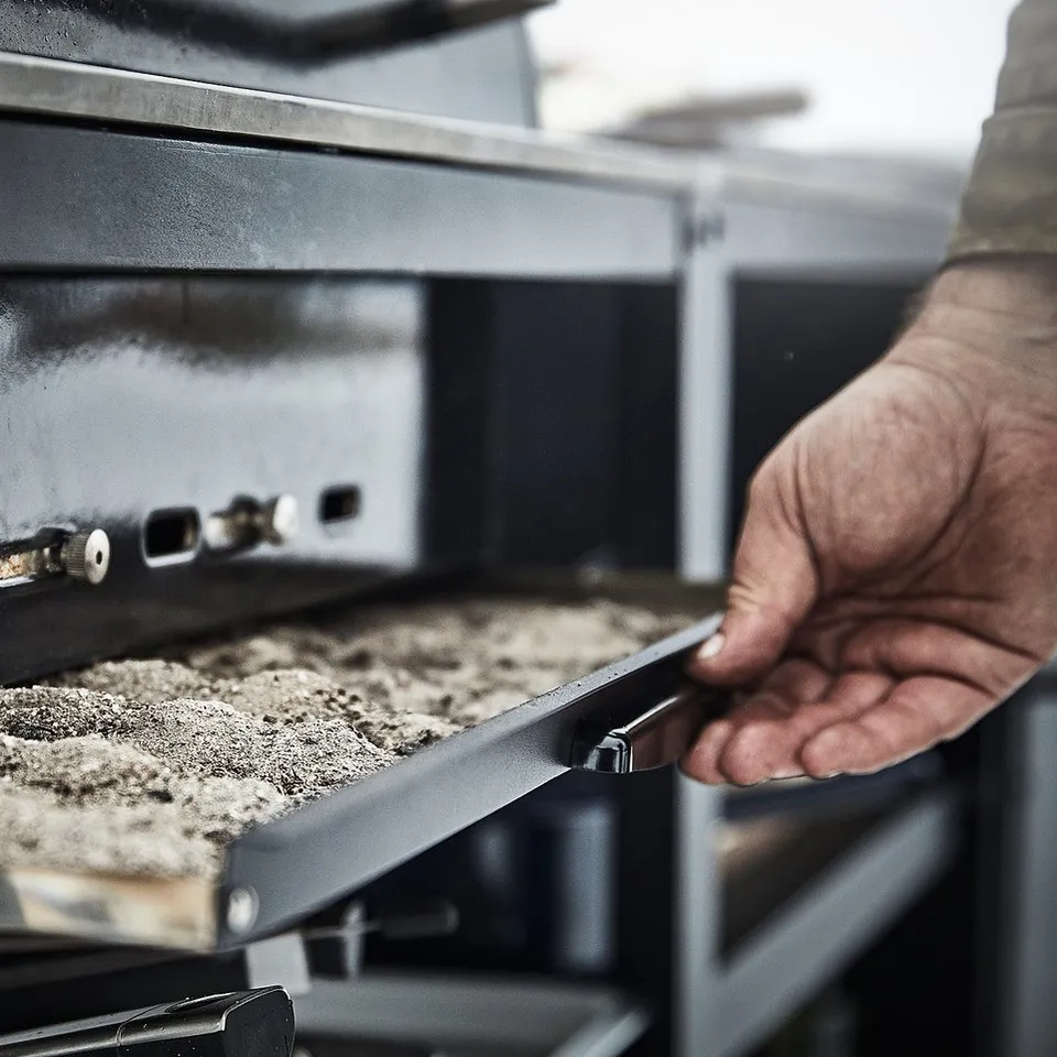A person opens a stainless steel charcoal grill, GRILLSKÄR, preparing for barbecue with coals inside.
