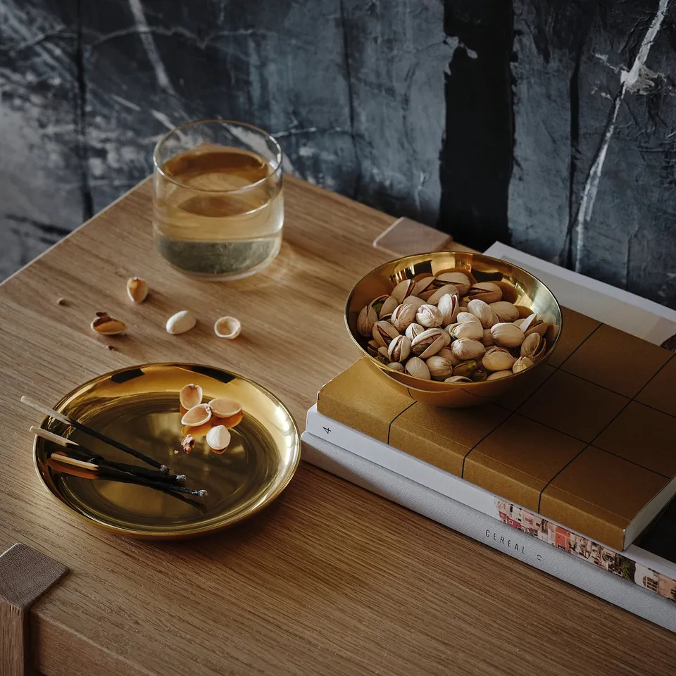 A wooden table holds a brass bowl filled with pistachios, a dish with matches and shells, and a cup of espresso. A book sits beneath the bowl.