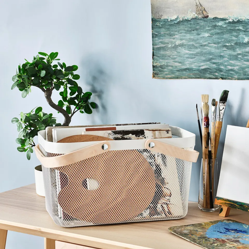 White RISATORP basket with handles on desk, holding papers, brushes, and a plant.