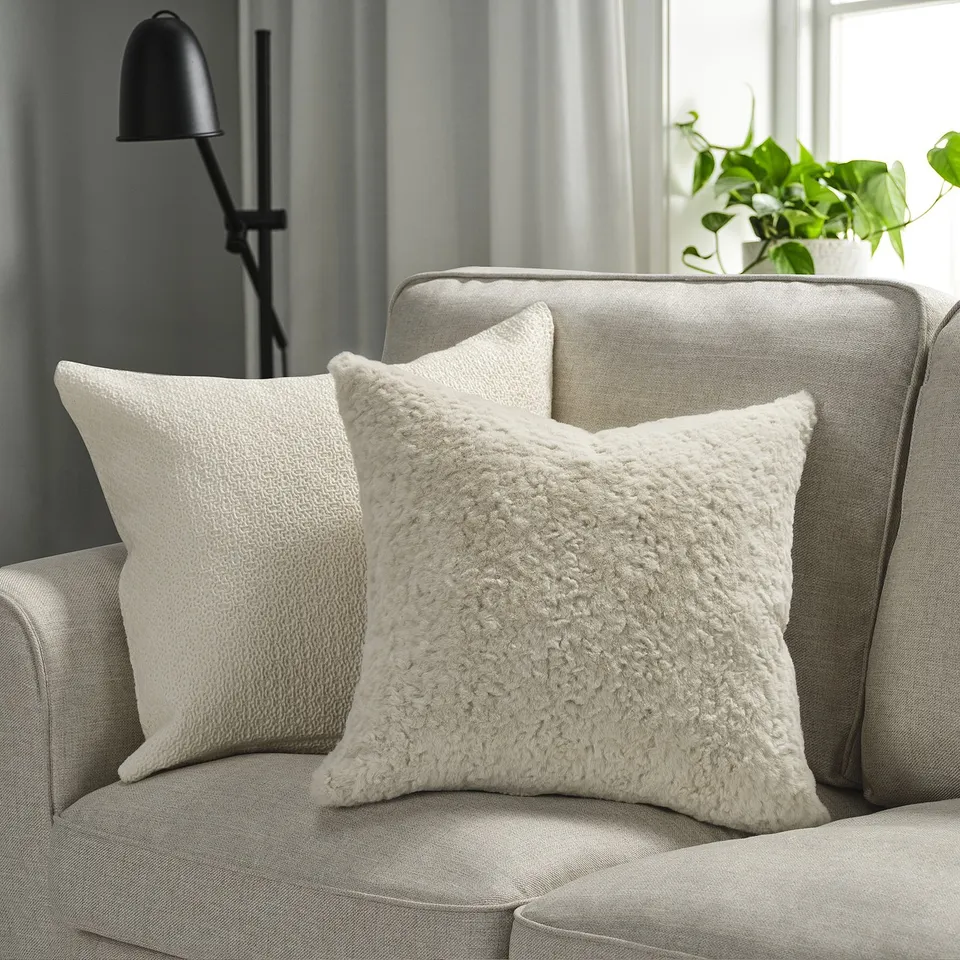 Two cream-colored, textured cushions on a grey sofa, one fluffy with a zip, beside a black floor lamp and a green plant.