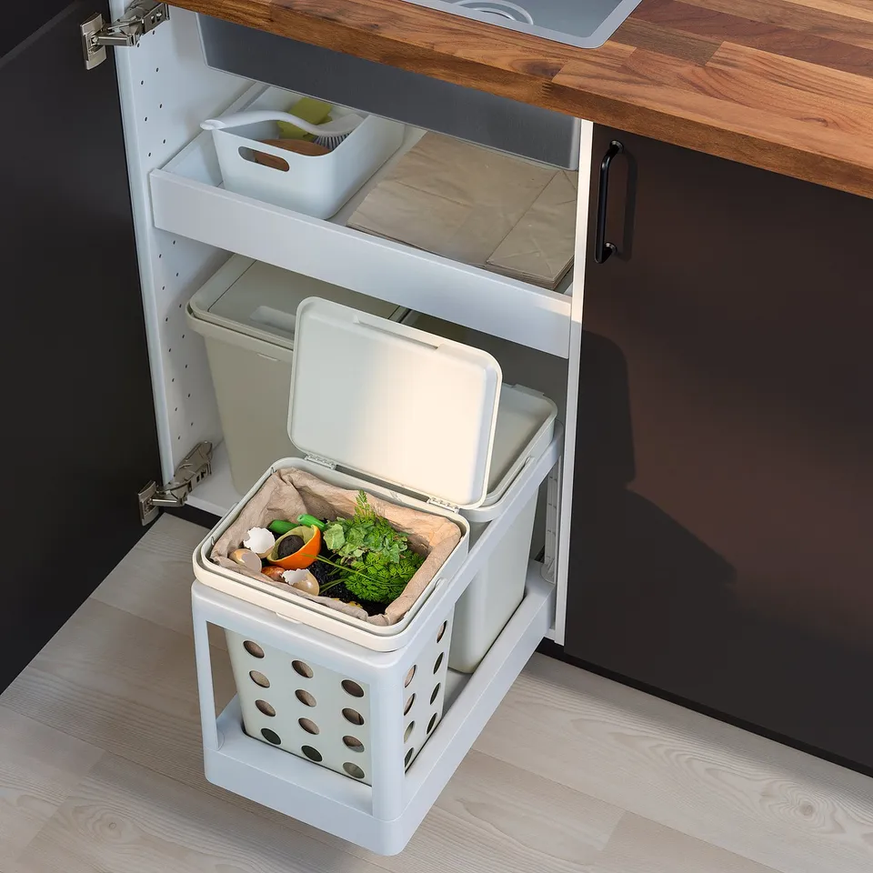 An open white HÅLLBAR compost bin under a kitchen cabinet, containing food waste. The bin has ventilation holes and a removable lid.