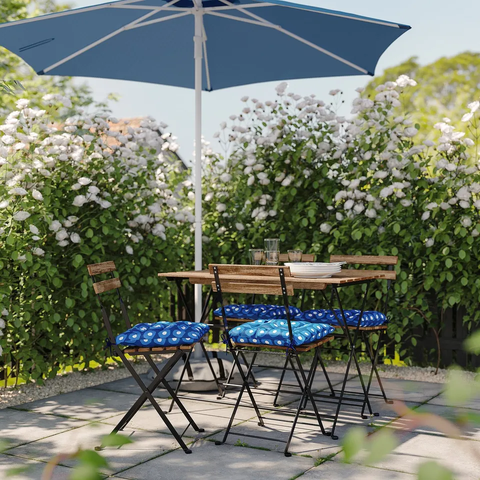 Small outdoor dining set with blue polka dot cushions, wooden table, and matching chairs beneath a large blue umbrella, surrounded by lush greenery.