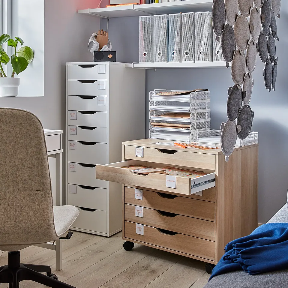 Office setup with light wood ALEX drawers, white shelves, desktop accessories, and hanging felt decorations. Chair, plant, and colourful wall.