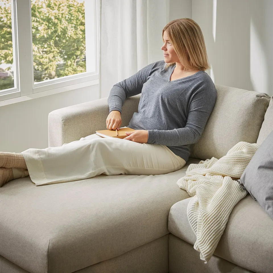 A person relaxes on a grey VIMLE sofa by a window, holding a book, with beige pillows and a blanket nearby.